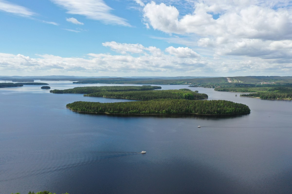 Low forested islands lying in wide, calm blue water, seen from above, with a distant treeline on the horizon.